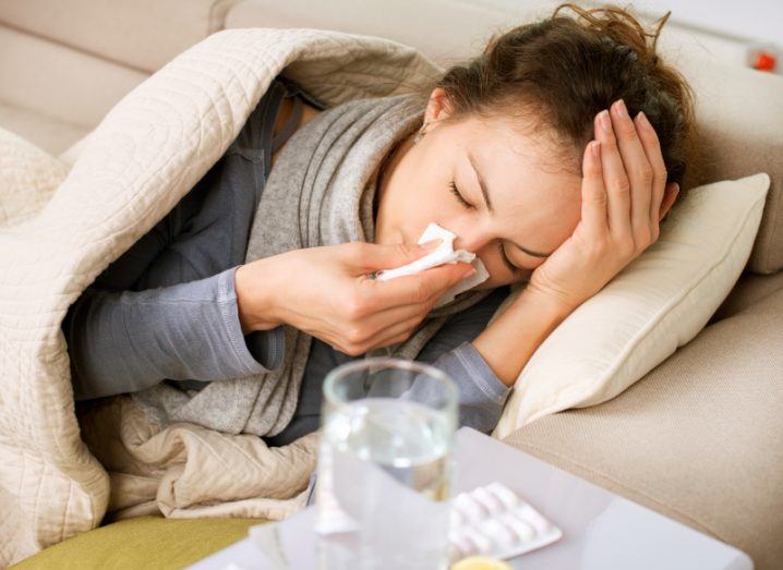 A woman lies on a couch wrapped in a blanked and blowing her nose. There is a blurred glass or water and a packet of tablets in the foreground to show she is sick.