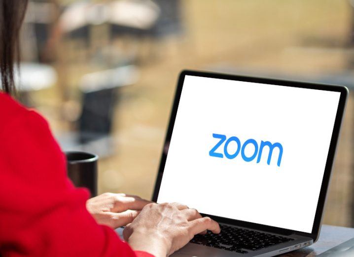 The blue Zoom logo on a laptop screen on a table with a person's hands on the keyboard and their arms in a red shirt visible. The office background is blurred.