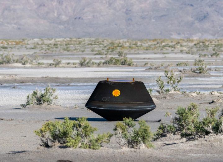 A black spacecraft capsule in the middle of a desert region, with green shrubs around it and large hills in the background.