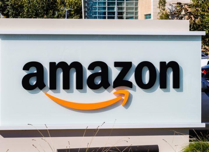 The Amazon company logo on a white sign in front of a building, with some trees visible in the background.