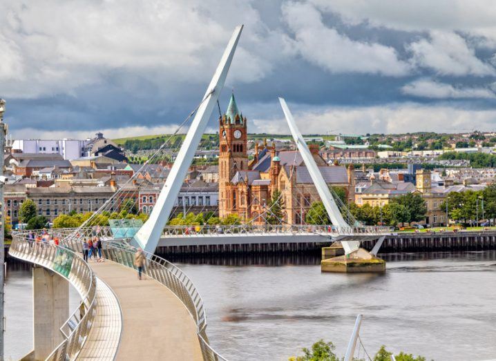 A bridge in Derry with buildings behind it and a cloudy sky in the background. A river is visible in the middle of the image.