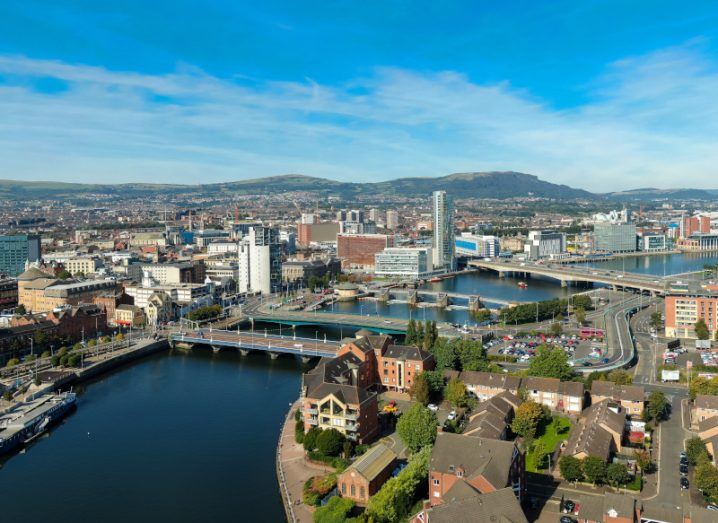 An aerial view of Belfast, the capital city of Northern Ireland. Buildings and a river are visible in the image, with a blue sky in the distance.