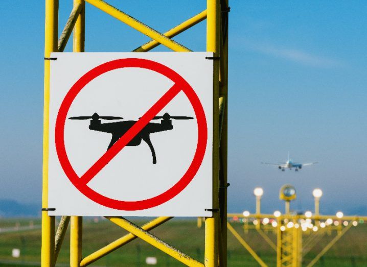 A sign on a yellow pillar, showing a drone with a red circle and line on it, signalling a location where drones are not allowed. A plane is visible in the background.
