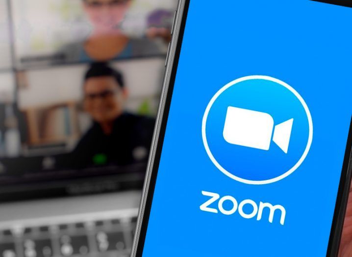 The Zoom logo on a smartphone screen, with a laptop screen in the background that has two people's faces on it.