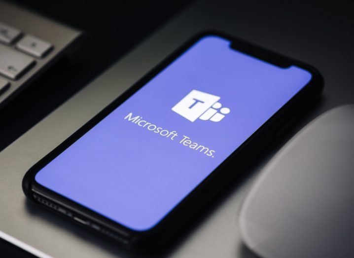 The Microsoft Teams logo on a smartphone screen. The phone is laying on a grey table next to a keyboard.