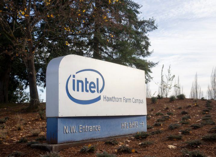 A sign in a brown field with the Intel logo on it, with trees and a grey sky in the background.