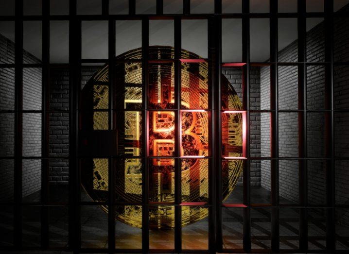 A bitcoin logo behind prison bars in a dark cell.