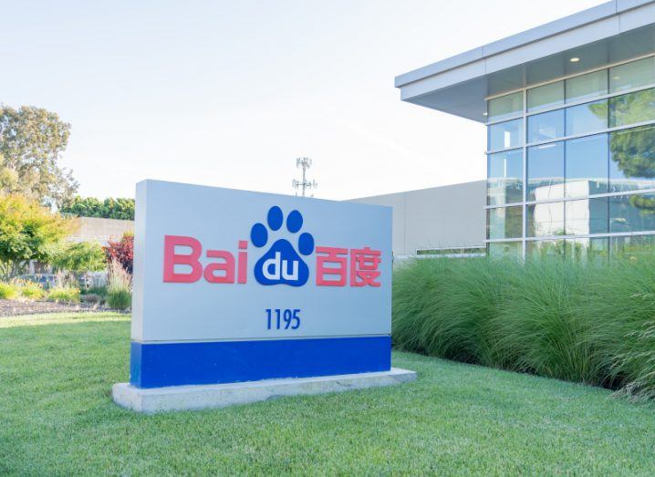 The Baidu company logo on a sign that is on a patch of green grass in front of hedges and a building.