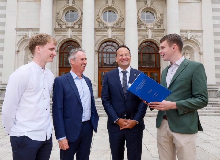 Four men standing together in front of a large building, with brown doors and circular windows. They are part of BT Ireland, the Irish government and BT Young Scientist winners.