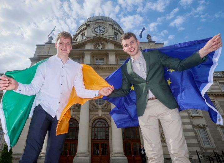 Two young men standing in front of a building, with one holding an Irish flag and the other holding an EU flag. They are Shane O'Connor and Liam Carew.