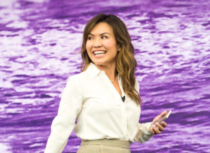 A woman smiles while looking over her right shoulder. She is standing in front of a purple digital conference screen and is wearing a mic on her shirt. She is Linh Lam.