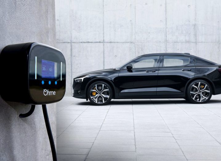 A black car in a white concrete room, with an Ohme smart charger attached to a wall.