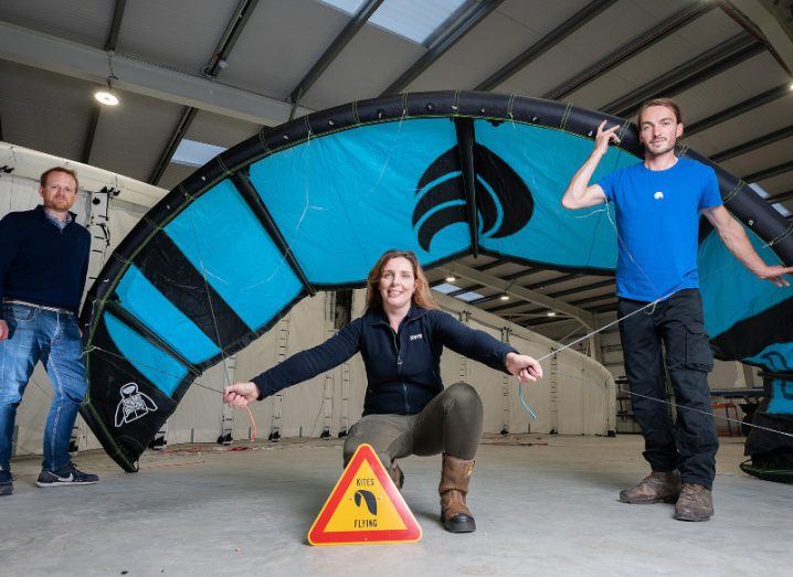 Two men and a woman standing around a large blue kite in a warehouse. The woman is on one knee while holding strings connected to the kite behind her.