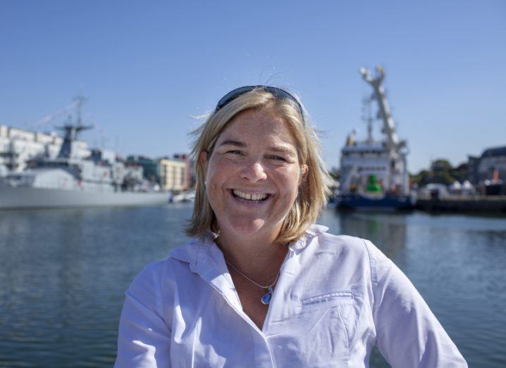A woman smiles at the camera. There is a dock behind her with a body of water. She is Vera Quinlan, a marine scientist at the Marine Institute.