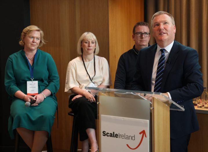 Minister Michael McGrath addresses a crowd from a podium at a Scale Ireland event with three people in the background.