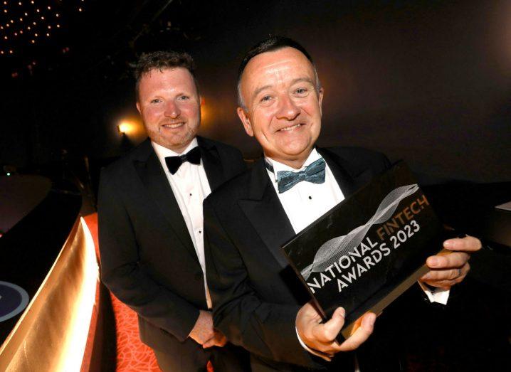 Colm Lyon holds his fintech award and smiles. There is a man smiling behind him and both of them are wearing suits with bowties.