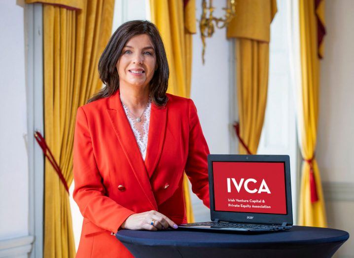 Headshot of Denise Sidhu, the new IVCA chair, wearing a red blazer and holding a laptop that has the IVCA logo on it.