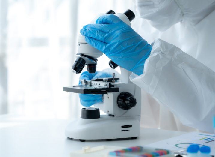 A researcher wearing lab overalls and blue surgical gloves looks into a microscope on top of a white lab desk.