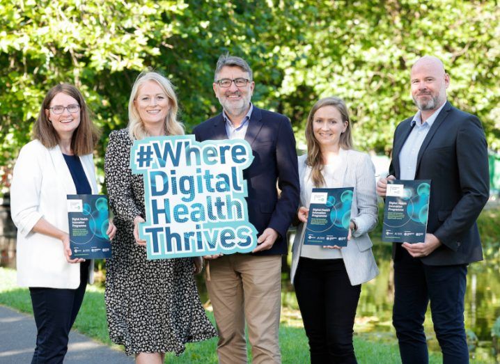 Three women and two men standing outside with some shrubbery behind them. Three of the people are holding booklets for the Digital Health Innovation programme, while two of the people are holding a sign with the hashtag "Where Digital Health Thrives".