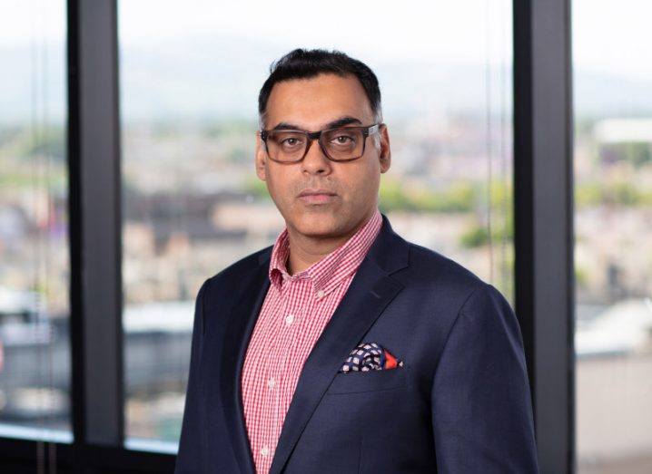 A man wearing a suit and glasses stands in front of a glass window. There is a blurred city landscape seen through the window. He is Puneet Kukreja, UK and Ireland cyber leader at EY.