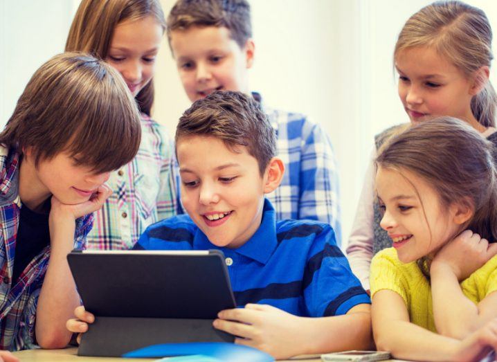 A group of young boys and girls gathered around one boy holding a smart device smiling at whatever is on screen.