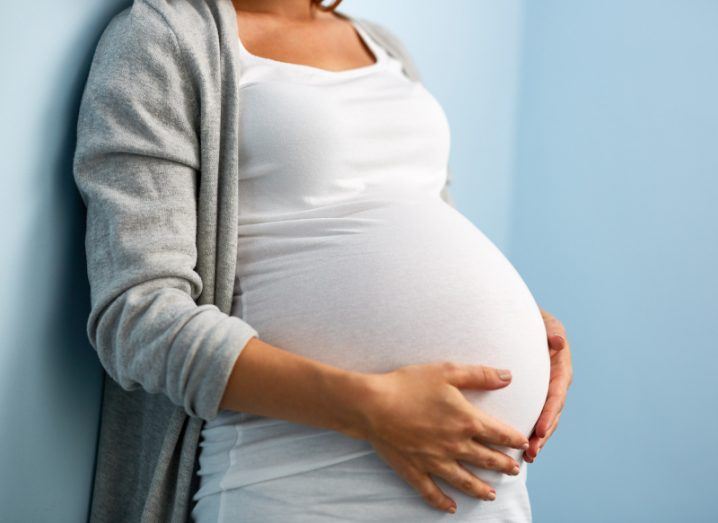 A close-up of a woman's pregnancy bump. Her hands are cradling the bump and she is wearing a white top and a grey cardigan, leaning against a blue wall.