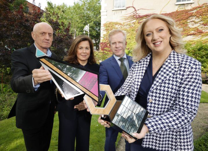 Two women and two men pose outside a building with trees and shrubs behind them. They are dressed in business suits. The woman on the right is Una Fitzpatrick and she holds an example of a Technology Ireland Industry Award.