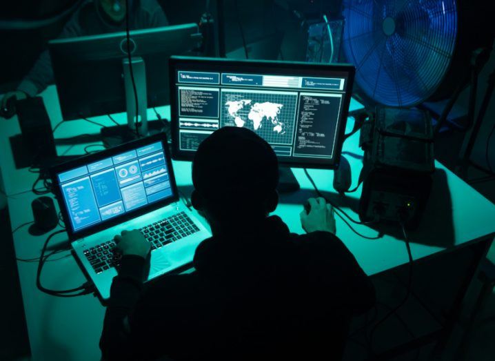 A person in a dark room, typing on two computers on a desk. Used to show the concept of cybercrime.