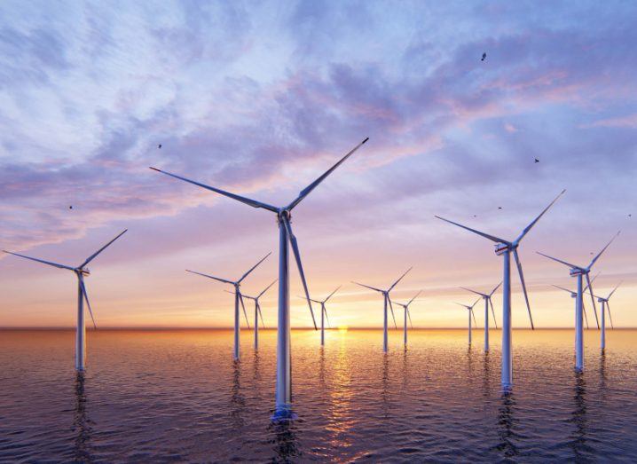 Multiple offshore wind turbines in the sea, with the sun setting in the background.