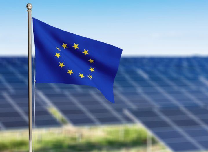 The EU flag in front of multiple solar panels, with a blue sky in the background.