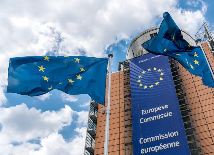 Two EU flags in front of a building which also has the EU logo on it, with clouds and a blue sky above the building.