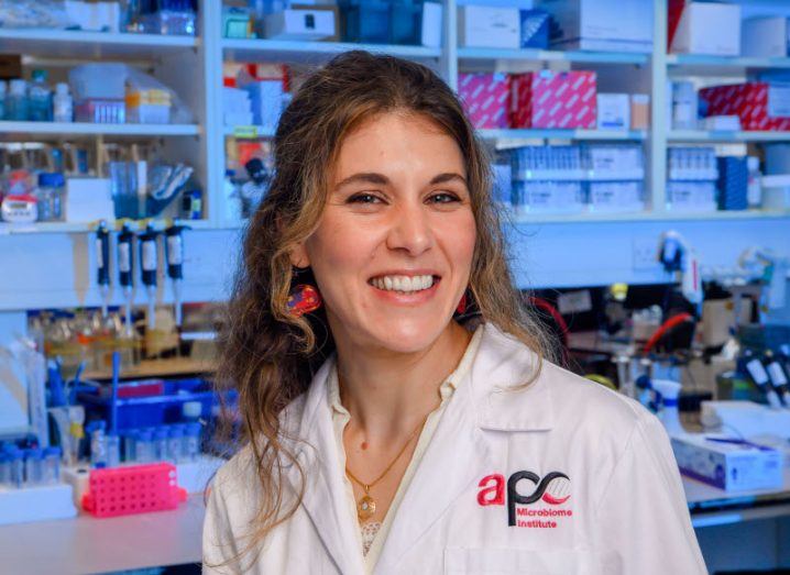 A woman in a lab setting, wearing a lab coat that has the APC Microbiome Ireland logo on it. She is Doctor María Rodríguez Aburto.