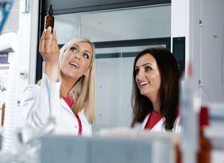 Shorla Oncology founders Orlaith Ryan and Sharon Cunningham stand together in a lab. Orlaith holds up an amber glass bottle with a pipette inside.