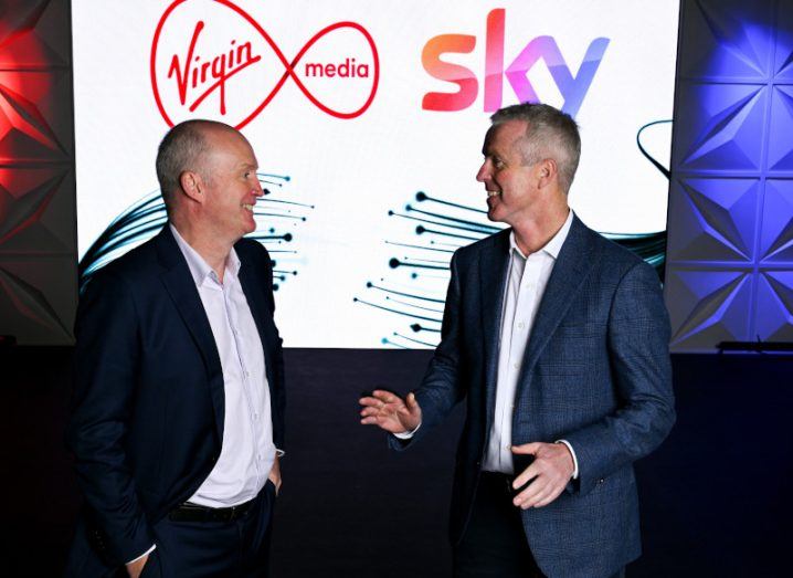 Two men in suits speaking to each other in front of a screen that has the Virgin Media and Sky logos on it.