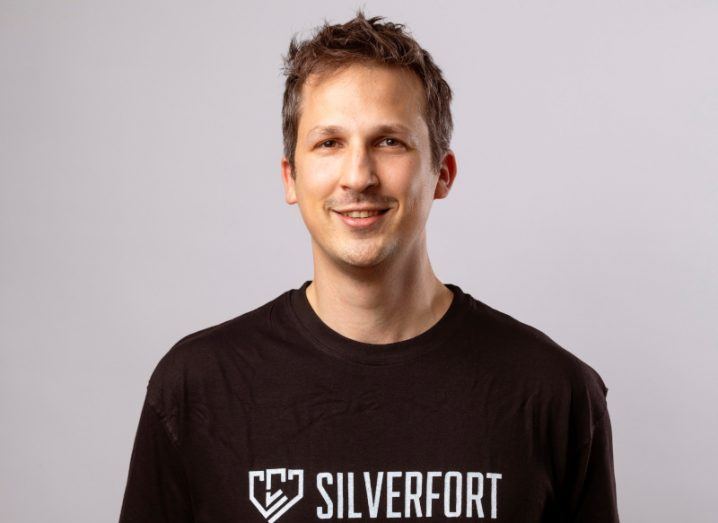 A man smiles at the camera in front of a grey background. He is wearing a dark shirt with the word 'Silverfort' on it in white writing. He is Yaron Kassner, co-founder and CTO of Silverfort.