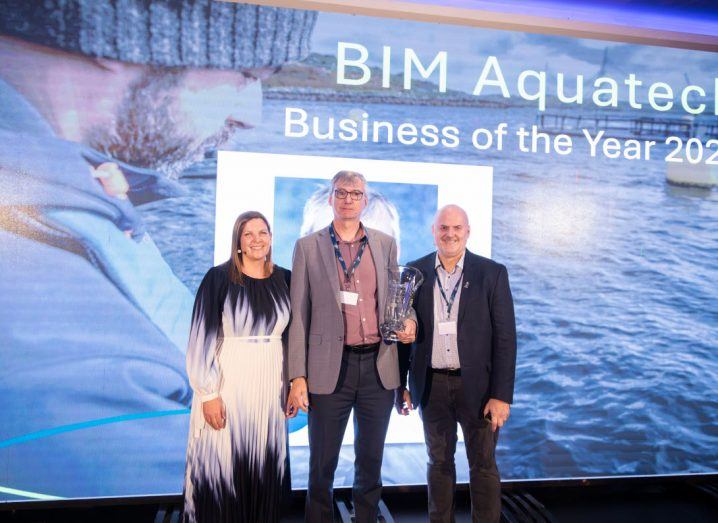 A woman and two men stand on a stage. The man in the middle is holding the BIM Aquatech Business of the Year award in his hand.