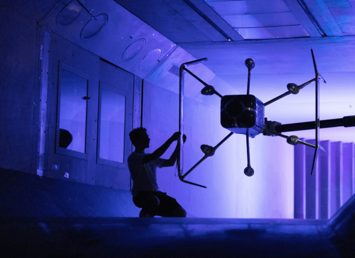 A dar, purple shaded image of a man kneeling down next to a large drone suspended from a low ceiling.