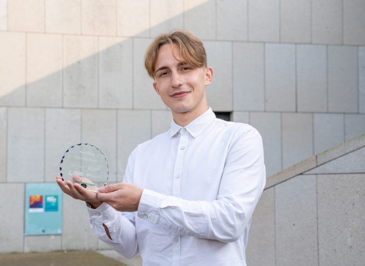 Snappie co-founder Mihael Melnic in a white shirt holds up a New Frontiers award for best pitch in front of a cream wall.