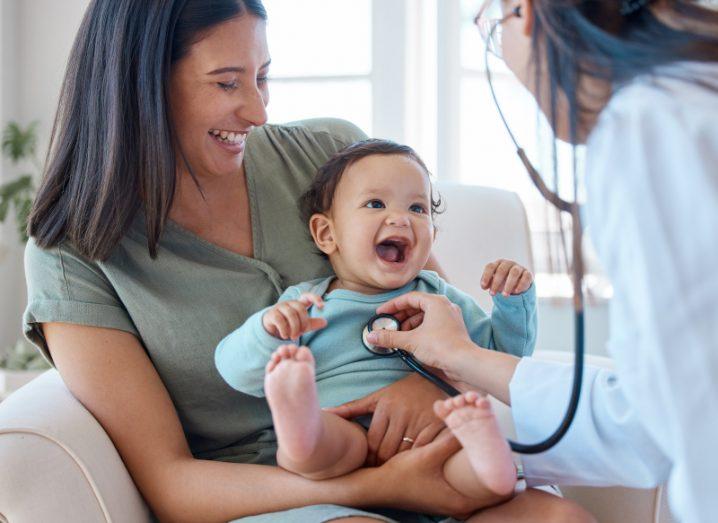 A woman holds a baby in her arms as she sits down. A doctor has a stethoscope placed on the baby's chest. They are all smiling and laughing.