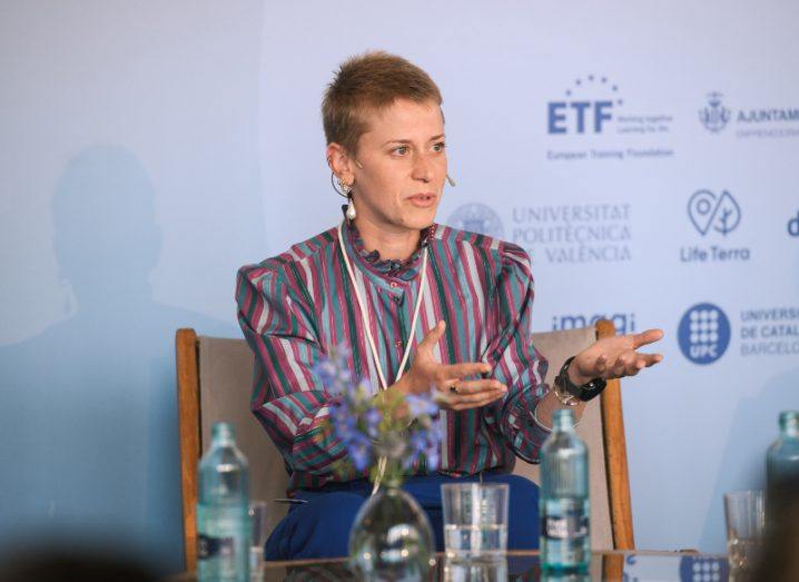 Cécile Deprez, aerospace researcher, sits speaking on a stage with a desk with a glass and bottle of water in front of her. She is talking. She wears a high-necked strpy blouse and has very short blonde hair. Her arms are outstretched, mid gesture.