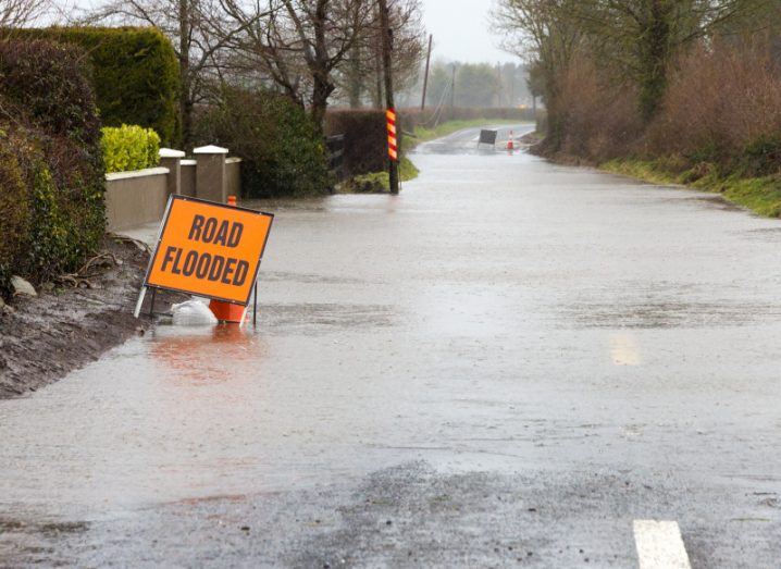 A road is flooded with a orange sign with the words 'road flooded' and a residential wall and trees and countryside visible.