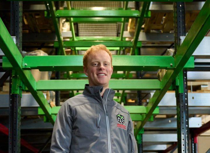 A man in a grey outfit standing in a warehouse in front of green panels. He is Sam Moffett.