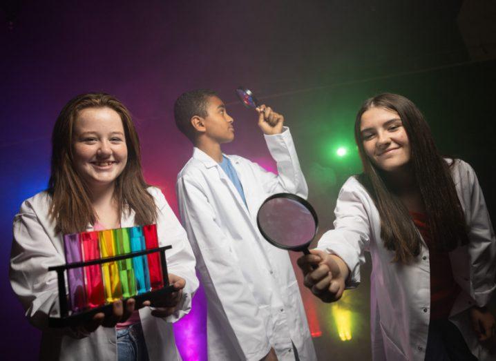 Three young students in white lab coats, in a dark room with different coloured lights in the background. One student is holding glass vials of different colours, while the two other students are holding magnifying glasses.