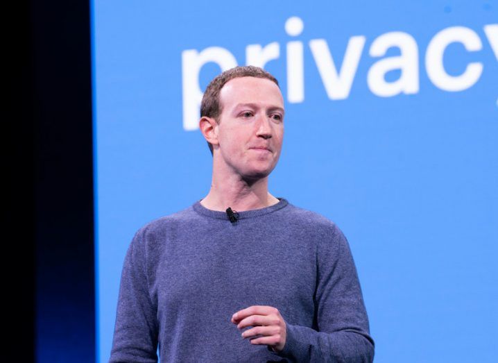Mark Zuckerberg wearing a blue jumper and standing on a stage, in front of a wall that has the word privacy written on it.