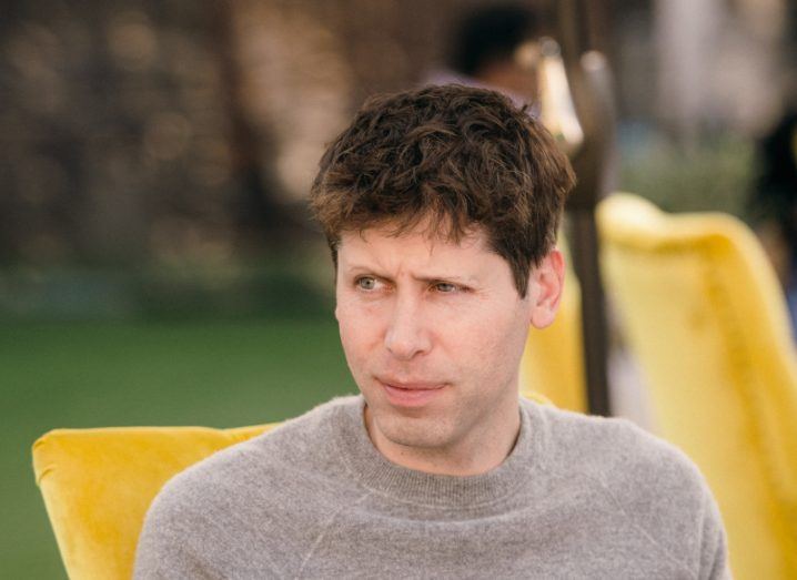 A man in a grey jumper sitting on a yellow chair. He is Sam Altman.