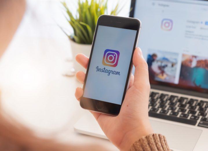 The Instagram logo on a smartphone screen, which is being held in a person's hand. A laptop is in the background which also has the Instagram logo on the screen.