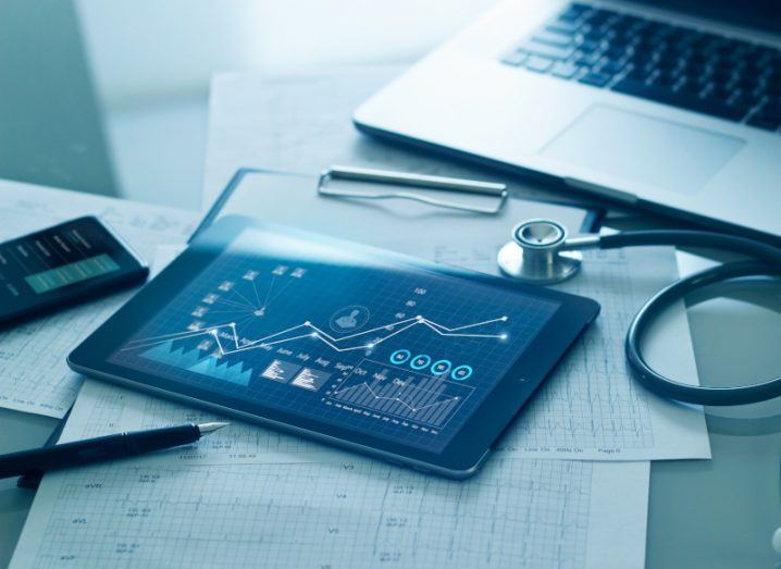 A table with a tablet laying on it and a graph on the screen. There are pieces of paper on the table, along with a laptop and a stethoscope.
