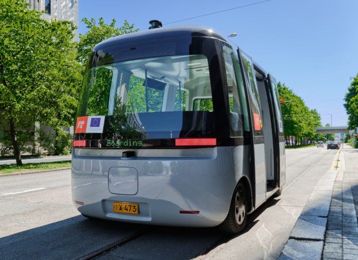 A self-driving vehicle in the middle of a road, with trees and buildings in the background.
