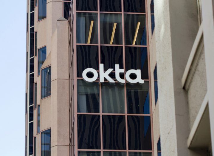 The Okta company logo on the side of a brown building, with windows visible on the side of the building and a grey sky on the left side of the image.