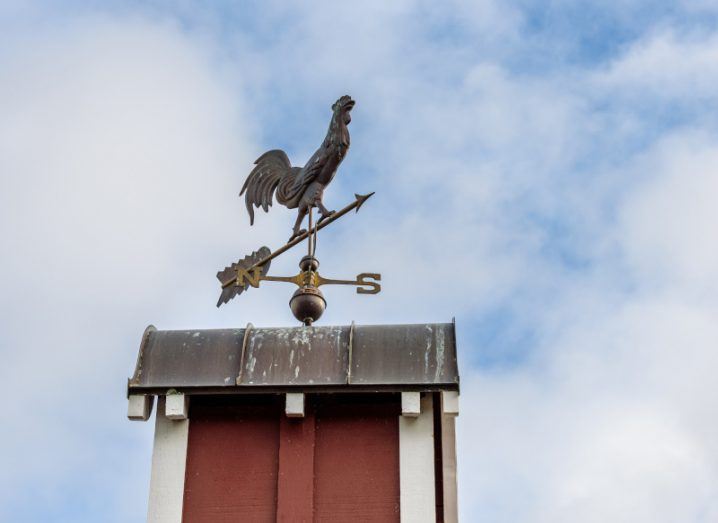 A weather vane on the top of a building, with an image of a grey rooster on top and an arrow pointing out, with a cloudy sky in the background. Used to represent weather prediction.